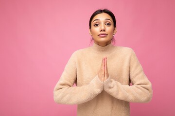 Portrait of young serious beautiful brunette woman with sincere emotions wearing casual beige jersey isolated over pink background with free space and keeping hands together asking for forgiveness