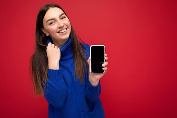 Closeup of delightful beautiful happy young brunette woman wearing casual blue sweater isolated over red background with empty space holding in hand mobile phone and showing smartphone with empty