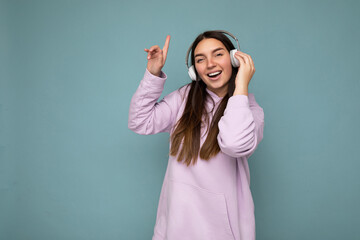 Attractive positive smiling young brunette woman wearing light purple hoodie isolated over blue...