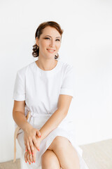 Portrait of a beautiful Caucasian medical girl in a white coat sitting on a stool on a white background. beauty salon.