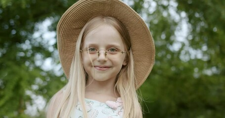 Cute little girl model with glasses and hat, smiling and looks in camera in park