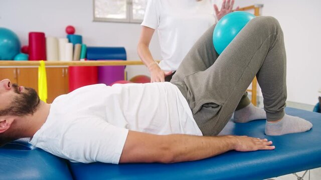 Handheld shot of a Caucasian man doing bridge and squeeze exercise, using fitness ball. Physiotherapy exercise with the ball between the knees for the inner thighs and good leg alignment.
