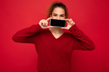 Closeup Photo of happy funny young female person wearing dark red sweater isolated over red background holding smartphone and showing mobile phone screen with copy space for cutout looking at camera
