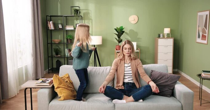 Caucasian beautiful young woman sitting on sofa practicing yoga, meditating with calm relaxed face while her small cute daughter kid girl jumping on couch in house. Relaxation. Motherhood concept
