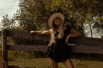 Gorgeous blond farm girl at the wood fence in a hat and short black dress posing