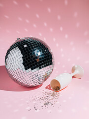 Shiny mirror disco ball with white champagne glass and silver confetti on pastel pink background....
