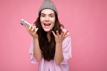 Naklejka premium Positive happy attractive young brunette woman wearing stylish pink shirt and grey hat isolated over pink background holding in hand and using mobile phone communicating and recording voice message