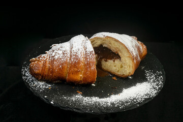 2 halves of a chocolate croissant, powdered with sugar, on a black stone tile, isolated on black background. 