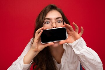 photo of shocked beautiful young brunette woman wearing white shirt and optical glasses isolated over red background holding in hand and showing mobile phone with empty screen for cutout looking at