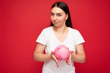 Fototapeta na wymiar Portrait photo of positive young beautiful attractive brunette woman with sincere emotions wearing casual white t-shirt isolated over red background with copy space and holding pink piggy box. Money