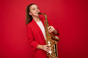 Fototapeta na wymiar Photo shot of pretty happy smiling brunette little girl wearing stylish red jacket standing isolated over red background wall playing saxophone looking to the side