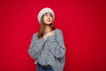 Portrait of positive happy young attractive dark blonde woman with sincere emotions wearing grey sweater and beige knitted hat isolated over red background with copy space and holding hands on chest