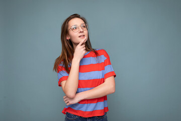 Photo of positive thoughtful young pretty brunette woman with sincere emotions wearing casual striped t-shirt and optical glasses isolated over blue background with copy space