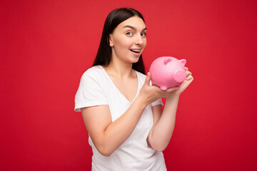 Obraz na płótnie Canvas Portrait photo of happy positive smiling young beautiful winsome brunette woman with sincere emotions wearing casual white t-shirt isolated over red background with copy space and holding pink pig