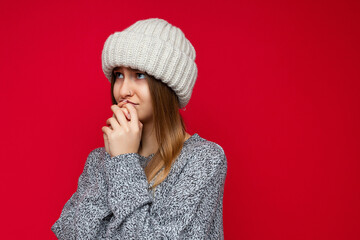 Closeup portrait of young winsome dark blonde woman with sincere emotions wearing grey sweater and beige knit hat isolated over red background with free space and praying with hands together asking