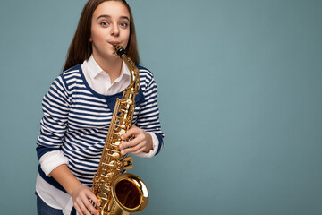 Fototapeta na wymiar Shot of attractive positive happy brunet little girl wearing stylish striped longsleeve standing isolated over blue background wall playing saxophone looking at camera