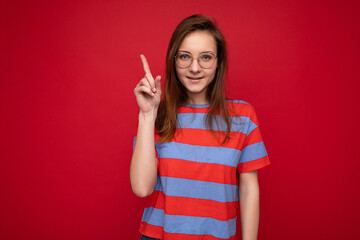 Positive happy young attractive brunette woman with sincere emotions wearing casual striped t-shirt and optical glasses isolated over red background with empty space and showing one finger