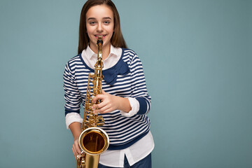 Beautiful happy smiling brunette little girl wearing striped longsleeve standing isolated over blue background wall playing saxophone looking at camera