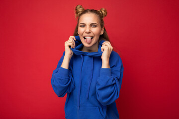 Young beautiful attractive pretty cool positive funny joyful blonde woman with two horns with sincere emotions wearing hipster blue hoodie isolated on red background with free space and showing tongue