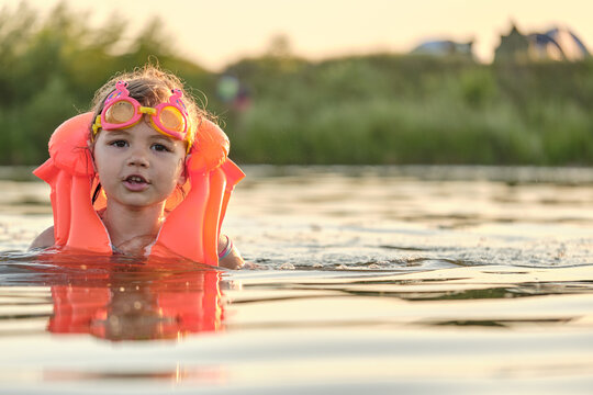 A little girl bathes in the water in the sunset light. Child playing on the beach during summer vacation. The child swims in a life jacket and goggles. Safety on the water.