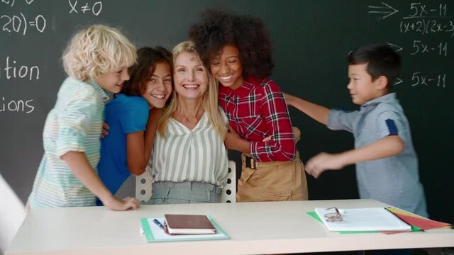Portrait of cheerful smiling teacher and happy diverse schoolchildren hugging on chalkboard blackboard background laughing looking at camera glad after school reopen on mathematics lesson.