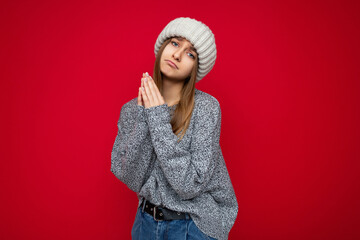 Portrait of young pretty dark blonde woman with sincere emotions wearing grey sweater and beige knitted hat isolated over red background with free space and praying with pray gesture