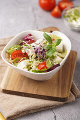 A small white salad bowl with microgreens, cherry tomatoes and fresh basil leaves on a wooden board on a grey concrete surface
