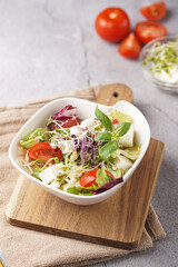 A small white salad bowl with microgreens, cherry tomatoes and fresh basil leaves on a wooden board on a grey concrete surface