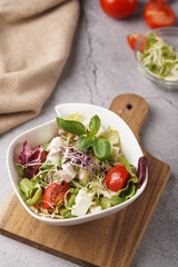 A small white salad bowl with microgreens, cherry tomatoes and fresh basil leaves on a wooden board on a grey concrete surface