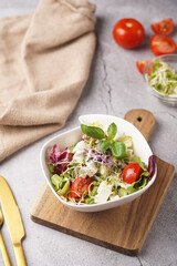 A small white salad bowl with microgreens, cherry tomatoes and fresh basil leaves on a wooden board on a grey concrete surface