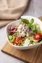 A small white salad bowl with microgreens, cherry tomatoes and fresh basil leaves on a wooden board on a grey concrete surface