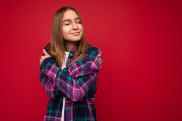 Fototapeta premium Photo of positive smiling cute happy young beautiful dark blonde woman with sincere emotions wearing hipster purple check shirt isolated over red background with copy space and hugging herself