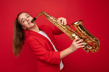 Fototapeta na wymiar Photo shot of beautiful happy smiling brunette little girl wearing stylish red jacket standing isolated over red background wall playing saxophone looking at camera