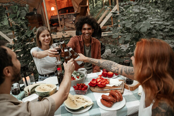 Young friends having dinner at glamping clincking glasses after sunset. Happy millennial gang camping at open air picnic under bulb lights. Spending time with friends outdoors, barbeque party