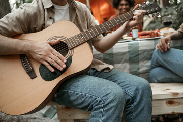 Obraz premium Faceless portrait of young man playing guitar hanging out traveling together with friends in forest glamping having fun, roasting sausages sitting at dinner table near tent during summer hanging out