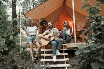 Obraz na płótnie Canvas Group of young friends traveling in glamping in the forest having fun playing guitar, mbira or kalimba and singing songs roasting sausages sitting at tent during summer vacation laughing hanging out