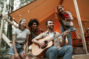 Fototapeta na wymiar Close up portrait of group of young friends hanging out traveling together in glamping in forest having fun playing guitar, making selfie, streaming to social media sitting in tent laughing