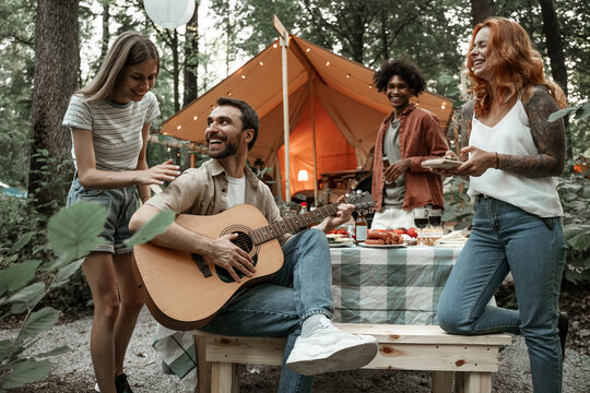Group of young friends traveling in glamping in the forest having fun playing guitar and mbira kalimba roasting sausages sitting at dinner table near tent during summer vacation laughing hanging out
