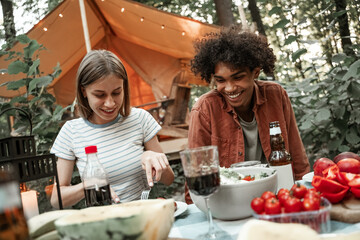 Young multiracial couple having dinner at glamping, laughing after sunset. Happy millennials camping at open air picnic under bulb lights. Spending time with friends outdoors, barbeque party