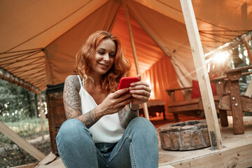 Red-haired smiling girl sits next to the tent and chatting messaging streaming on the smartphone, posting, liking in social media. Travel outside city in the woods. Camping.