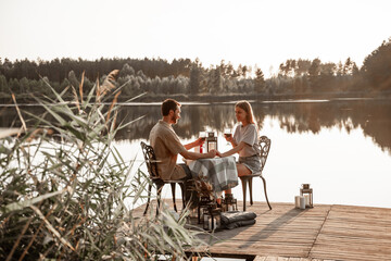Young couple sitting at the table spend time together on wooden pier on forest lake celebrating anniversary, holding hands. Love is in the air, love story concept. Romantic date on lake with candles.