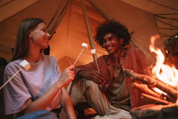 Young smiling couple roasting marshmallows on skewers over fire pit at campsite, looking at each...