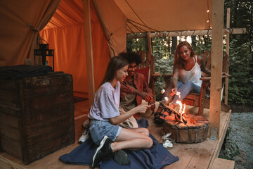 Group of young smiling people roasting marshmallows on skewers over fire pit at campsite, enjoying outdoor glamping holiday with friends togetherness reopen after pandemic lockdown.