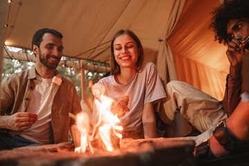 Group of young smiling people roasting marshmallows on skewers over fire pit at campsite, enjoying outdoor glamping holiday with friends togetherness reopen after pandemic lockdown