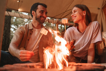 Young smiling couple roasting marshmallows on skewers over fire pit at campsite, looking at each other with love, enjoying outdoor glamping holiday togetherness reopen after pandemic lockdown