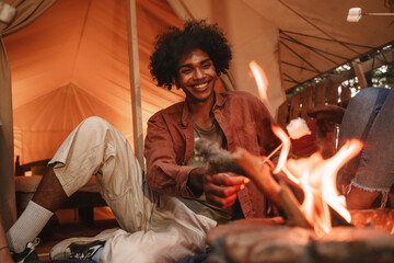 Hansome young curly african man roasting marshmallows on skewers over fire pit at campsite,...