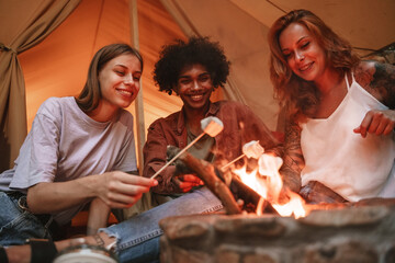 Group of young smiling people roasting marshmallows on skewers over fire pit at campsite, enjoying...