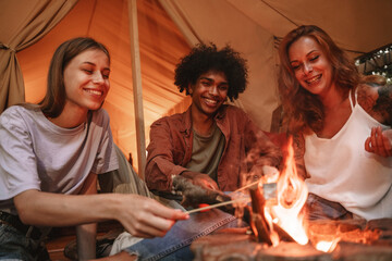 Group of young smiling people roasting marshmallows on skewers over fire pit at campsite, enjoying...