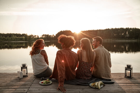 Group of friends having fun on picnic near a lake, sitting on wooden pier eating and drinking wine, beer, cider. Smiling young people having party celebration outdoors during sunset in countryside