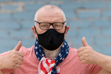 an elderly man with a gray beard and glasses tied an American flag bandana and a black mask. He's...